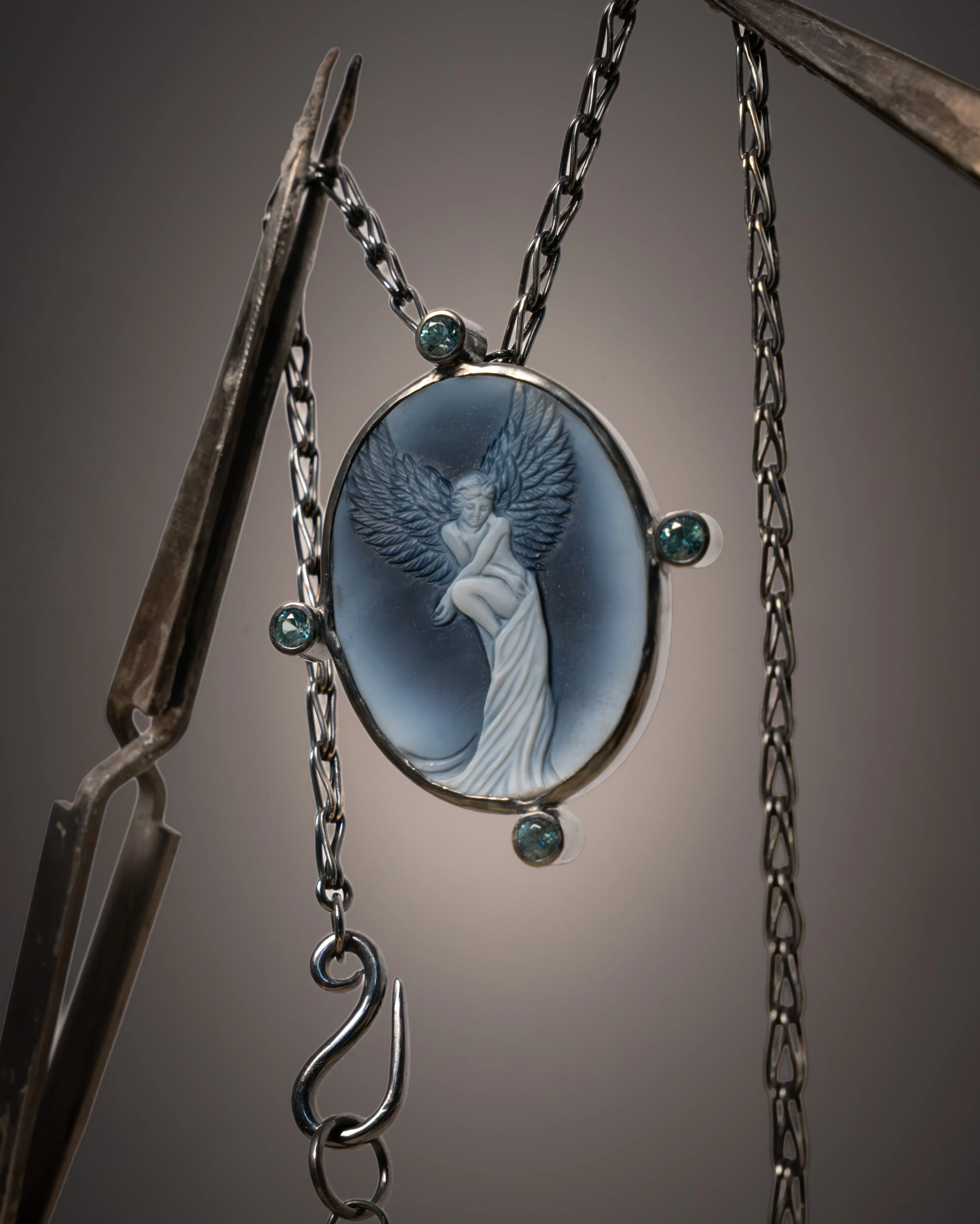 A winged cameo pendant held between two bench clamps in the workshop, its hand-forged chain hanging loose.