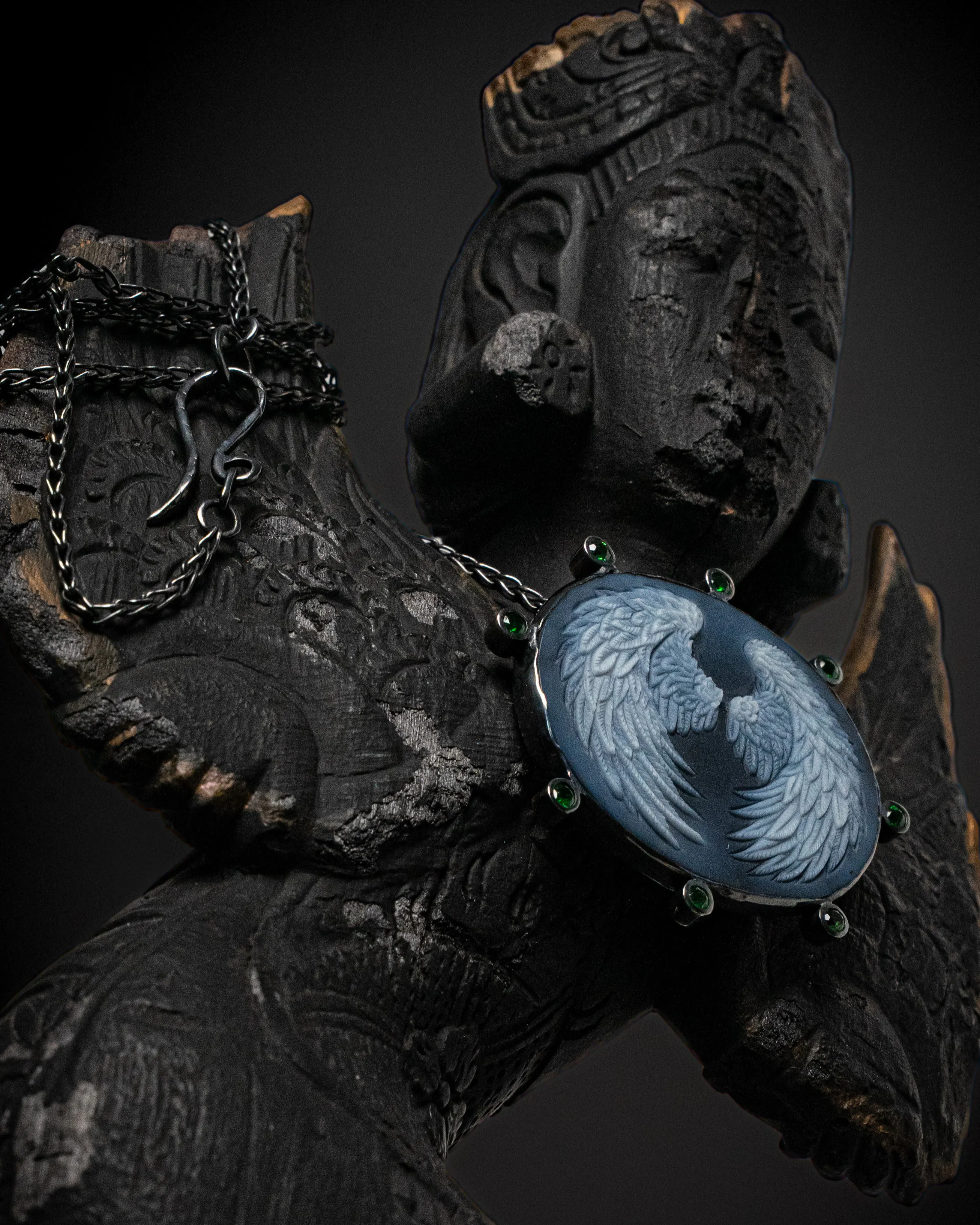 A winged cameo pendant in blue agate, ringed with green stones, resting against a carved and charred wooden statue.