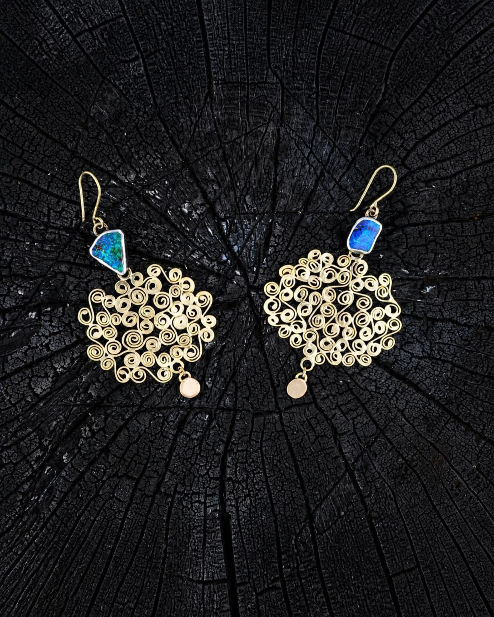 A pair of gold filigree earrings coiled from wire, set with opal, on a cross-section of burnt wood.