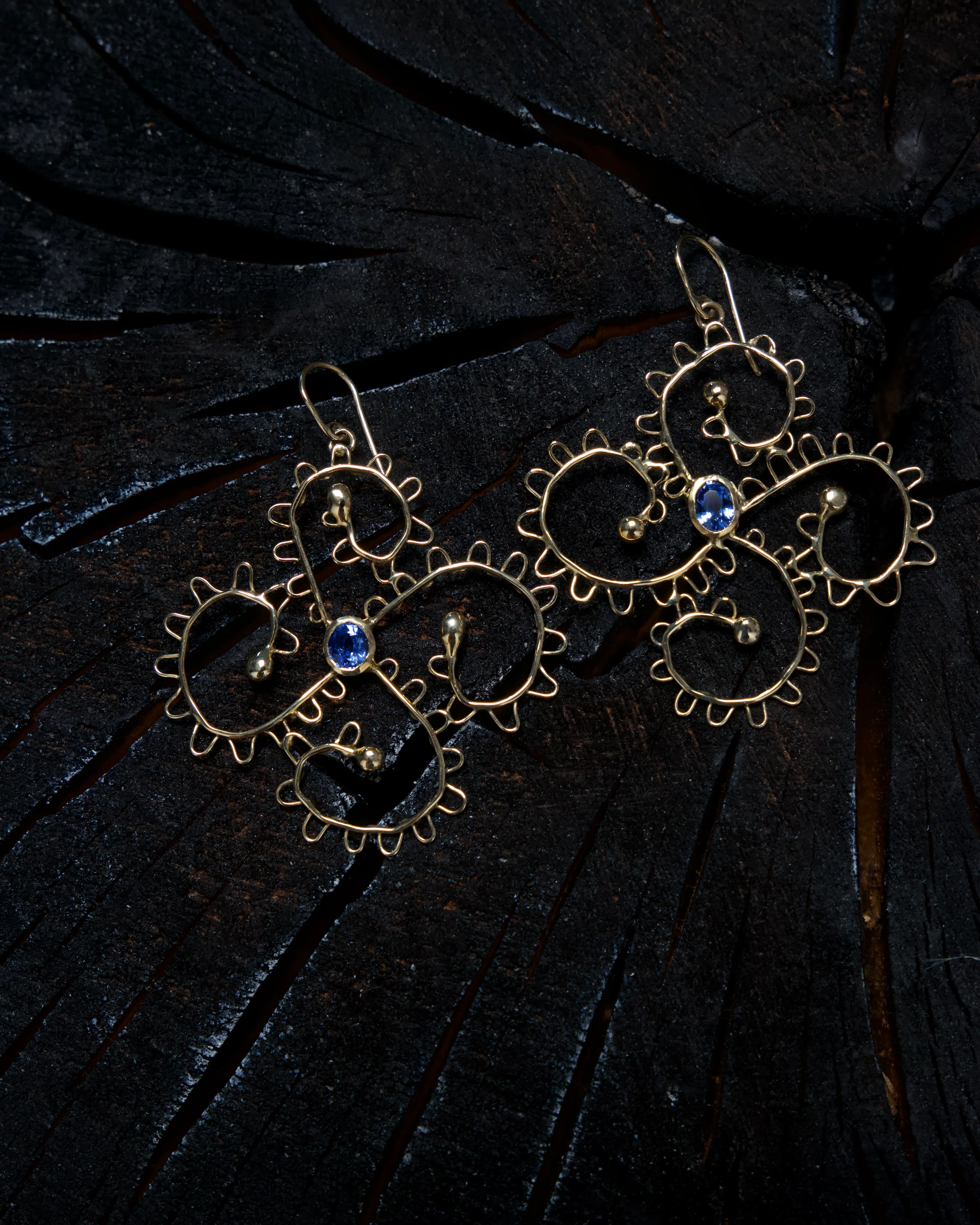A pair of gold wire earrings bent into open scrolls, each set with an oval blue stone, on charred wood.