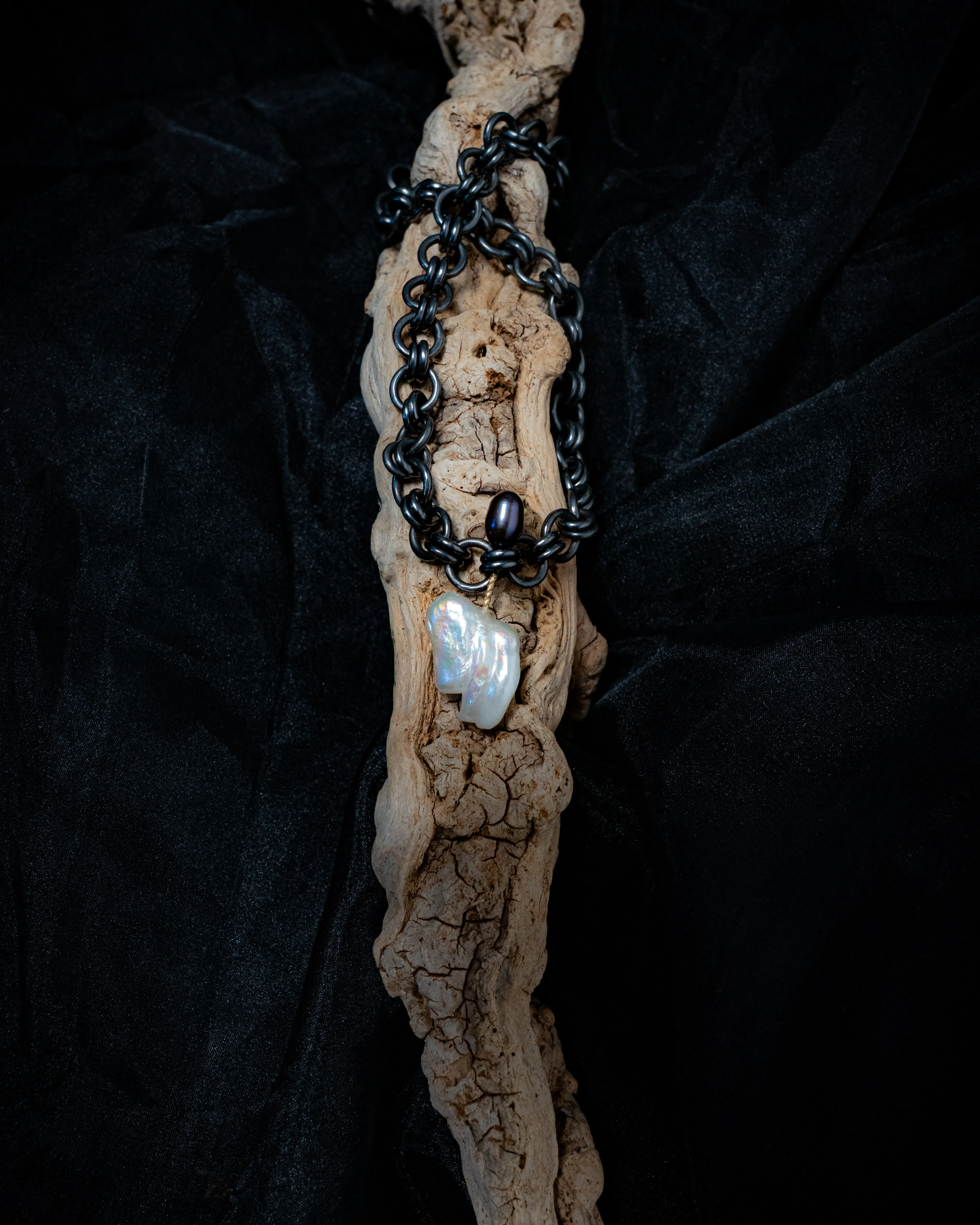 A blackened hand-forged chain and a single baroque pearl draped over pale driftwood.