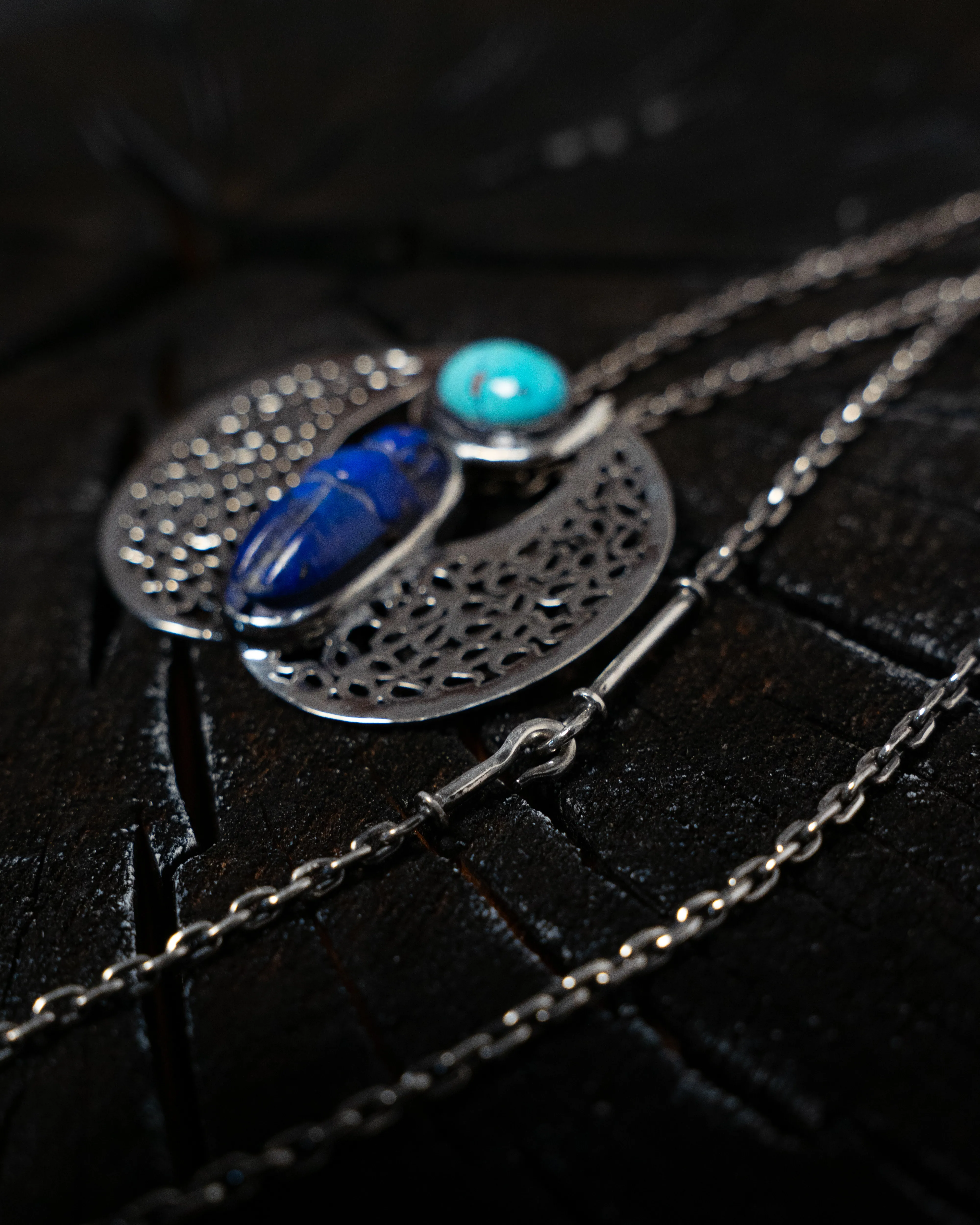 A pierced silver pendant set with lapis and turquoise, laid on charred wood.